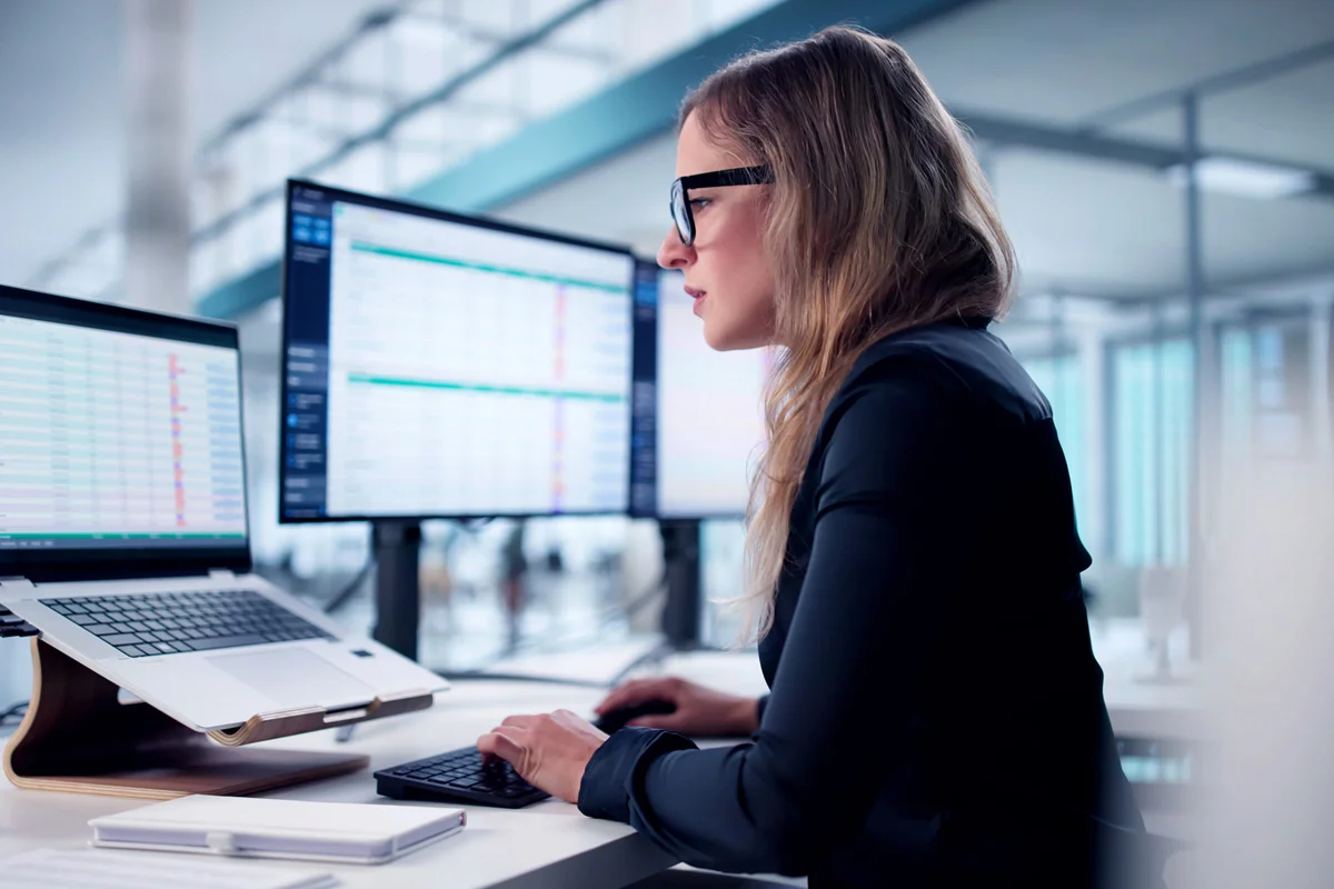 Woman in glasses at a desk looking at a laptop and monitor