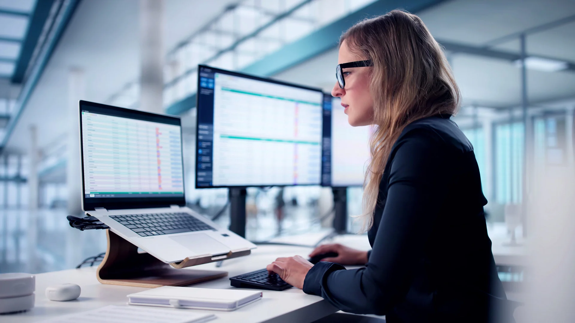 Woman in glasses at a desk looking at a laptop and monitor